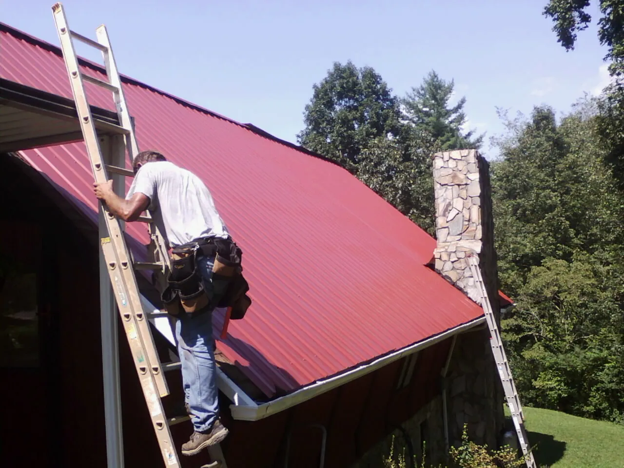 Roofer on a ladder beside a red metal roof and stone chimney — Soffit & Fascia Repair by Roofing Brothers in Nokesville